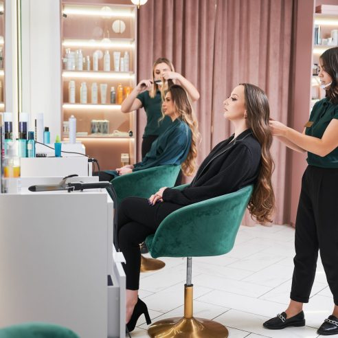 Professional hairdresser doing hairstyle for woman in beauty salon. Female hairstylist curling client hair with curling iron device while colleague combing woman hair.